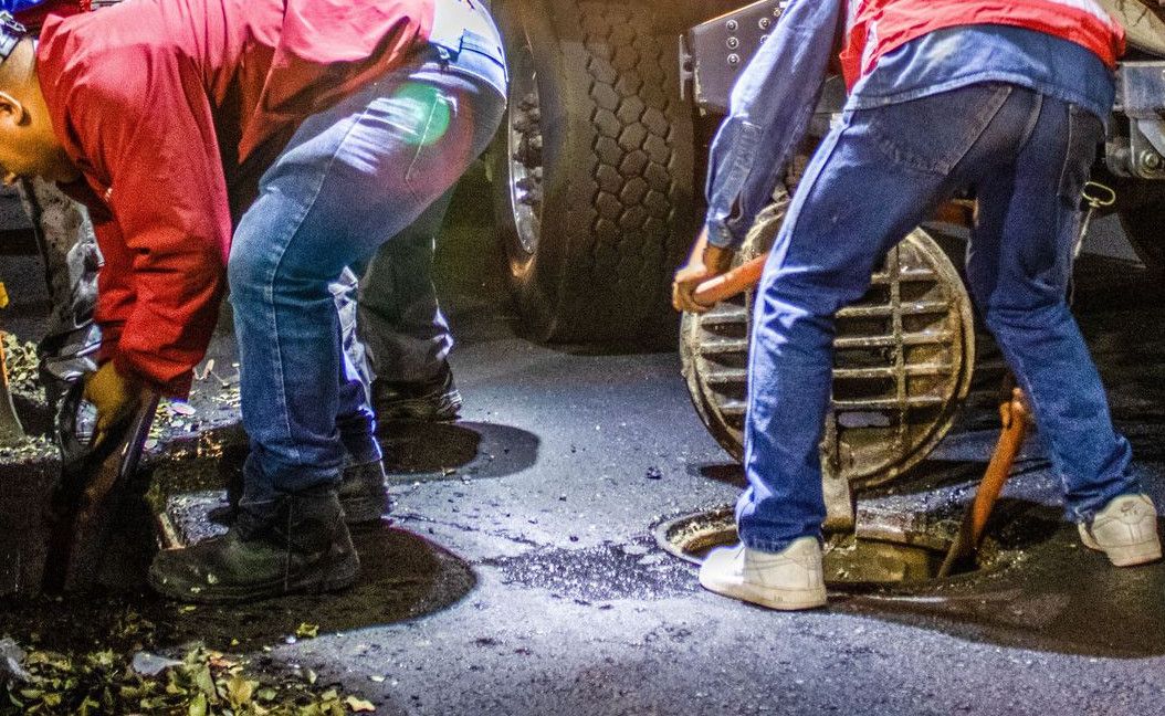 Street workers cleaning drainage at night with machinery under a bridge.